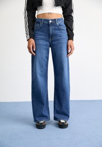 Jean en denim bleu à jambes larges et taille haute, avec cinq poches et des coutures contrastées. Porté avec des baskets noires et marron.