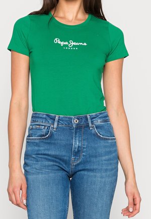 Kobieta ubrana w zielony t-shirt Pepe Jeans London i niebieskie jeansy z wysokim stanem, stoi na jednolitym jasnym tle.