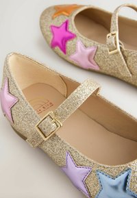 Ballerines dorées scintillantes avec une bride, ornées d'appliqués d'étoiles colorées en rose, violet, lilas et orange. Doublure intérieure lisse, forme classique.