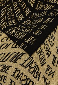 Tecido tricotado em preto e dourado com texto repetido em diferentes fontes e tamanhos. A textura é suave, com um leve brilho metálico.