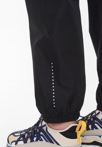 Černé sportovní kalhoty s elastickými manžetami, na boku s vertikálním řetězcem reflexních teček. Spárované se sneakers v béžové a modré barvě.