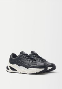 Sneakers in pelle nera con finitura testurizzata, forma arrotondata, suola intermedia bianca e suola in gomma nera. Presentano lacci e un logo sulla linguetta.