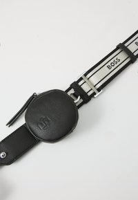 Bolso de cintura negro de cuero con un diseño redondeado, cuenta con un cierre de cremallera, unido a una correa de rayas en blanco y negro con la marca "BOSS".