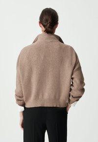 Pull beige à côtes avec un col montant et une coupe ample, doté de manches larges et d'un ourlet court, porté par-dessus une chemise blanche à col.