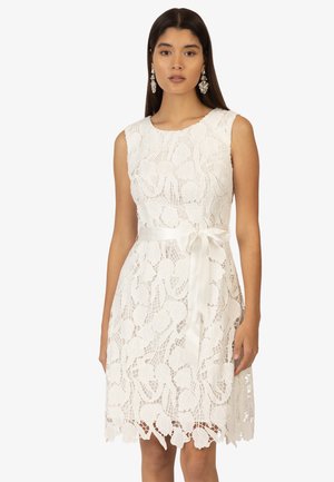 Robe blanche en dentelle avec des motifs floraux, sans manches, et une ceinture en satin à la taille. Longueur genou avec un ourlet festonné.