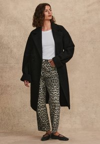 Schwarzer Wollmantel mit breitem Kragen, kombiniert mit hochtaillierten Jeans im Leopardenmuster und schwarzen Slippern. Glatte Textur.