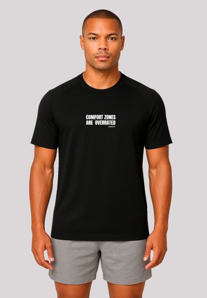 Athletischer Mann in schwarzem T-Shirt mit dem Aufdruck "COMFORT ZONES ARE OVERRATED LEAVE IT!" und grauen Shorts vor einfarbigem Hintergrund.