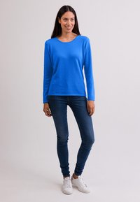 Maglione blu a maniche lunghe con scollatura rotonda, realizzato in materiale morbido, abbinato a jeans aderenti e sneaker bianche.