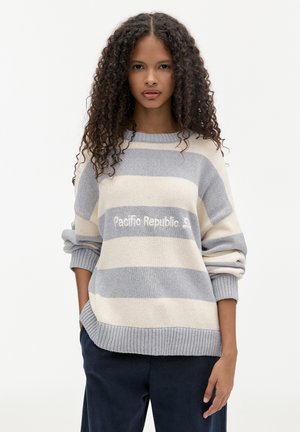 Pull rayé en gris clair et crème, avec une coupe décontractée, des poignets côtelés et le texte "Pacific Republic" sur le devant. Texture de tissu douce.