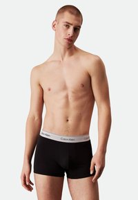 Black cotton boxer shorts with a gray waistband featuring the "Calvin Klein" logo, model standing with a neutral expression and bare torso.