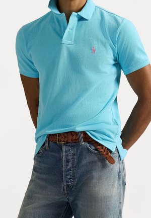 Homme portant un polo bleu clair avec un logo rose, ceinture en cuir marron tressée et jean bleu délavé, debout les mains dans le dos.