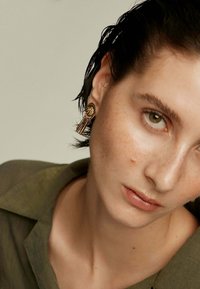 Pendientes en tono dorado con base circular que presenta patrones grabados y flecos de metal colgantes. El modelo tiene el cabello corto y oscuro, y un maquillaje neutro.