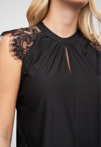 Le haut noir présente des manches en dentelle transparente et un décolleté en forme de trou de serrure. Tissu lisse avec un léger fronce aux épaules.