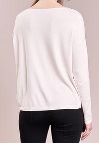 Haut léger à manches longues blanches avec un large col, coupe légèrement oversize et texture lisse, associé à un pantalon noir ajusté.