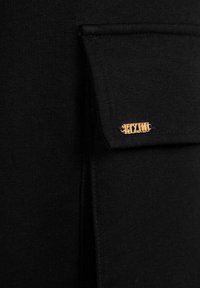 Czarna tkanina z kieszenią z złożonym klapkiem, podkreślona złotym detalem z logo "BIZUU". Tekstura wydaje się gładka i dziana.