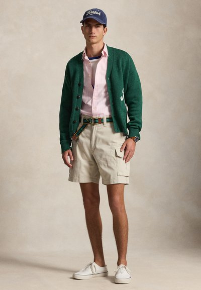 Mladý muž v zelenom kardigáne, ružovej pruhovanej košeli, béžových cargo kraťasoch, námorníckom modrom šiltovke, pruhovanom opasku a bielych teniskách stojí na jednolitej hladkej stene.