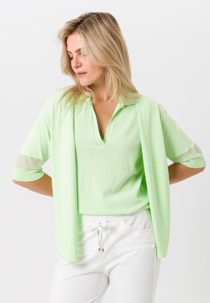 Haut vert clair à manches courtes avec un col en V, présentant des accents transparents sur les manches et une coupe ample, associé à un pantalon blanc.