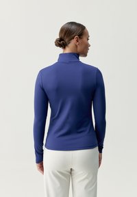 Långärmad, höghalsskjorta i djupblått stretchtyg, figursydd design, slät textur, i kombination med ljusa byxor, sedd bakifrån.