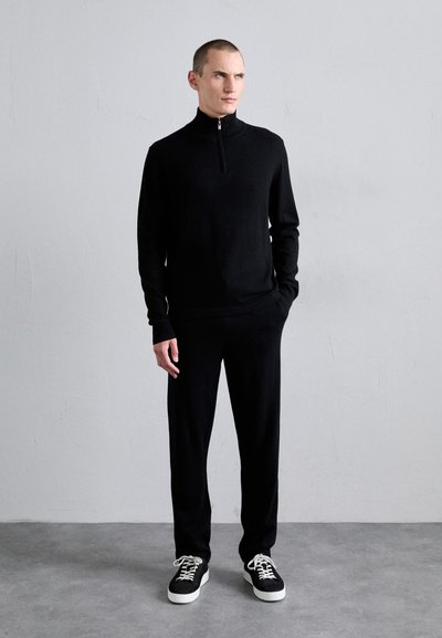 Black knitted zip-neck sweater paired with matching black trousers. Model wears black and white sneakers. Simple, streamlined design.