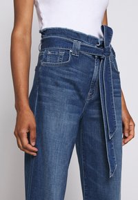 Denimowe spodnie z wysokim stanem, z postrzępionym paskiem, wiązanym w talii i klasycznym ciemnoniebieskim odcieniem. Posiadają boczne kieszenie i luźny krój.