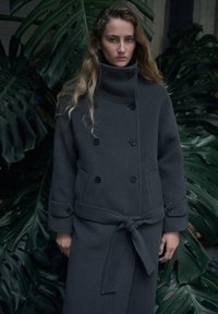 Cappotto doppiopetto grigio scuro con collo alto, tasche laterali e cintura in vita annodata, realizzato in un tessuto morbido e strutturato.