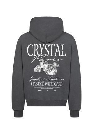Grijze hoodie met een grote witte afbeelding op de achterkant met "CRYSTAL Paris," diamanten en wasinstructies in cursieve en vetgedrukte lettertypes.