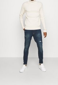 Un suéter de cuello alto blanco con canalé, jeans oscuros ajustados con detalles desgastados y zapatillas blancas. Atuendo moderno y casual.