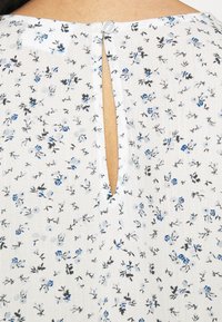 Kukallinen kangas, jossa on pieniä sinisiä ja mustia kukkakuoseja valkoisella taustalla. Ominaisuuksiin kuuluu avainaukko ja nappisulku.