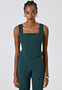 Top senza maniche color teal con scollatura quadrata e design strutturato, realizzato in tessuto morbido. Indossato con un bottom aderente coordinato. Accessori dorati.
