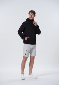 Schwarze leichte Jacke mit Kapuze, Reißverschluss vorne und Seitentaschen, kombiniert mit hellgrauen Cargo-Shorts mit schwarzen Akzenten. Weiße Sneakers.