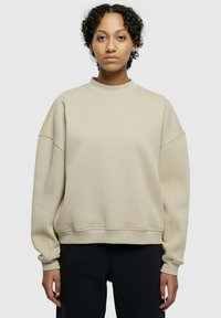 Beige sweatshirt med høy hals, fallende skuldrer, ribbestrikkede mansjetter og en myk tekstur. Har en avslappet passform og minimale sømmer.