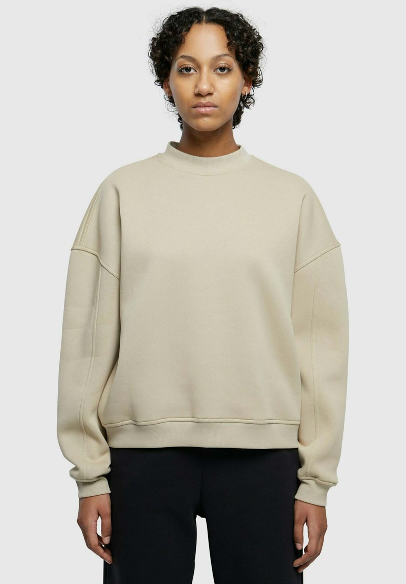 Felpa beige con collo alto, spalle cadenti, polsini a coste e una texture morbida. Presenta una vestibilità rilassata e cuciture minime.