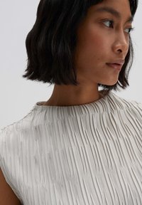 Pleated cream dress with a high neckline. Smooth texture with vertical ridges, showcased on a model turned slightly to the side.