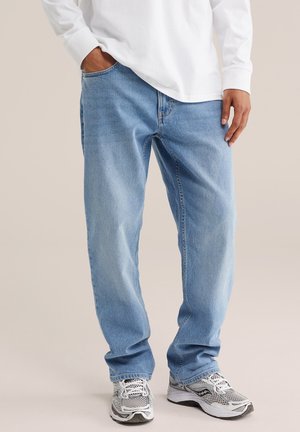Personne portant un jean ample bleu clair, une chemise blanche à manches longues et des chaussures de course blanc-gris, avec une main dans la poche.