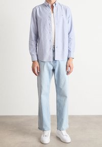 Man draagt een lichtblauw overhemd over een witte tanktop, lichtblauwe jeans met rechte pijpen en witte sneakers, staand op een grijze vloer.