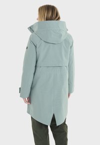 Veste imperméable vert clair avec capuche, dotée d'une ventilation à l'arrière et de poignets ajustables. Texture lisse et design arrivant aux genoux.
