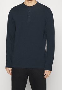 Man som bär en marinblå långärmad henley-tröja med tre knappar, ihop med svarta byxor, står mot en enkel vit bakgrund.