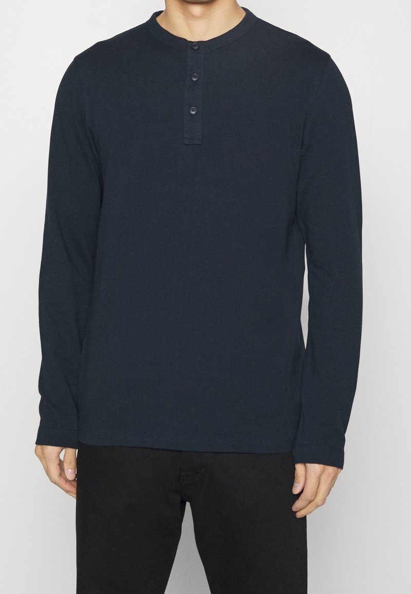 Man som bär en marinblå långärmad henley-tröja med tre knappar, ihop med svarta byxor, står mot en enkel vit bakgrund.