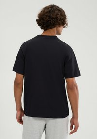 Schwarzes Baumwoll-T-Shirt mit kurzen Ärmeln, Rundhalsausschnitt und lockerer Passform, von hinten gezeigt, kombiniert mit hellgrauen Hosen.