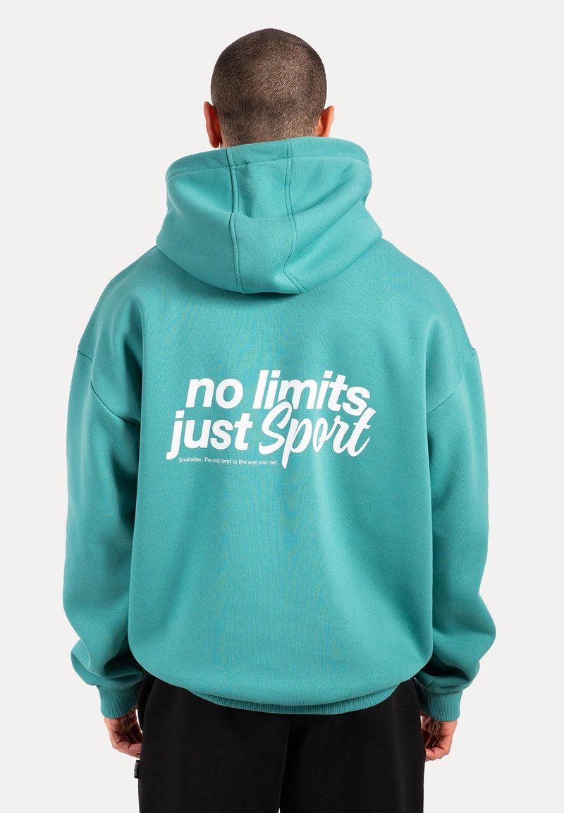 Homme portant un sweat à capuche bleu sarcelle avec un texte blanc "no limits just Sport" dans le dos, debout dos à un fond uni.