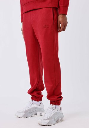 Pantaloni da tuta rossi con una texture liscia, caratterizzati da un piccolo logo sulla coscia, cintura elastica e polsini. Abbinati a scarpe da corsa bianche.