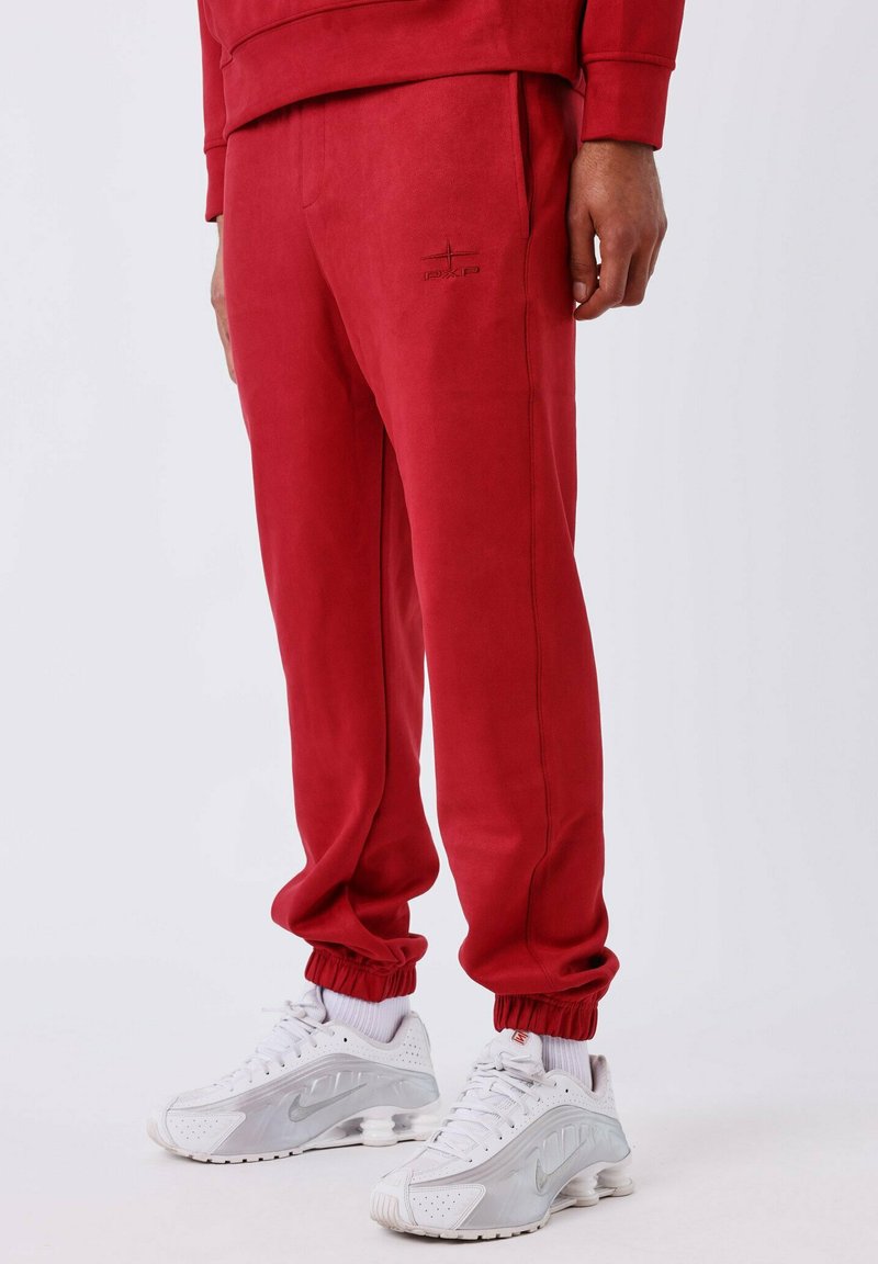 Pantalones de chándal rojos con una textura suave, que presentan un pequeño logo en el muslo, cinturilla elástica y puños. Combinados con zapatillas de running blancas.