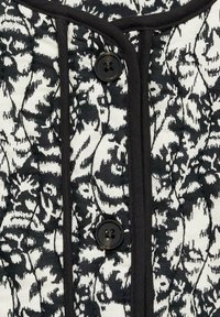 Zwart-wit gequilt textiel met een bloemenpatroon, omrand met zwarte rand. Twee zwarte knopen zijn gecentreerd langs de sluiting.