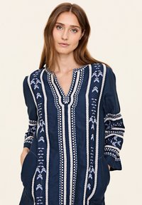 Tunic bleumarin cu broderie albă și albastră, decolteu în V, mâneci lungi și două buzunare laterale. Prezintă modele geometrice și o margine texturată.