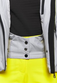 Chemise noire rentrée dans un pantalon jaune vif avec une veste gris clair partiellement ouverte, montrant des boutons noirs sur le revers intérieur de la veste.