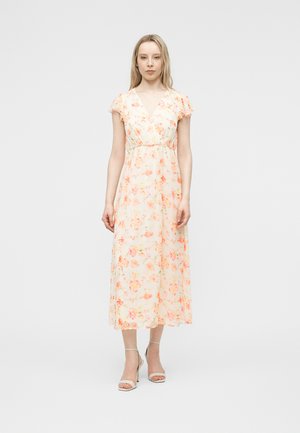 Frau steht in einem hellpfirsichfarbenen Midikleid mit Blumenmuster, mit flatternden Ärmeln und Knöchelriemchen-Pumps vor einem schlichten grauen Hintergrund.