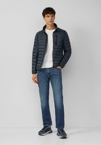 Granatowa pikowana kurtka z wysokim kołnierzem, z zamykaną na zamek kieszenią na piersi, noszona na białej koszuli, do tego niebieskie dżinsy i szare trampki.