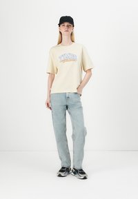 Lichtgele katoenen t-shirt met "TIMBS" graphic, gecombineerd met lichtblauwe jeans en een zwarte pet. Model draagt grijs en zwarte sneakers.