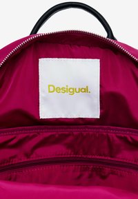 Intérieur d'un sac à dos fuchsia avec une poignée noire, comportant une étiquette blanche avec le texte jaune "Desigual" et une poche zippée. Texture en nylon lisse.