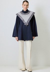 Tunic albastru marin cu guler din satin gri, având un design în formă de V și detalii cu funda la talie, asortată cu pantaloni evazați de culoare crem.